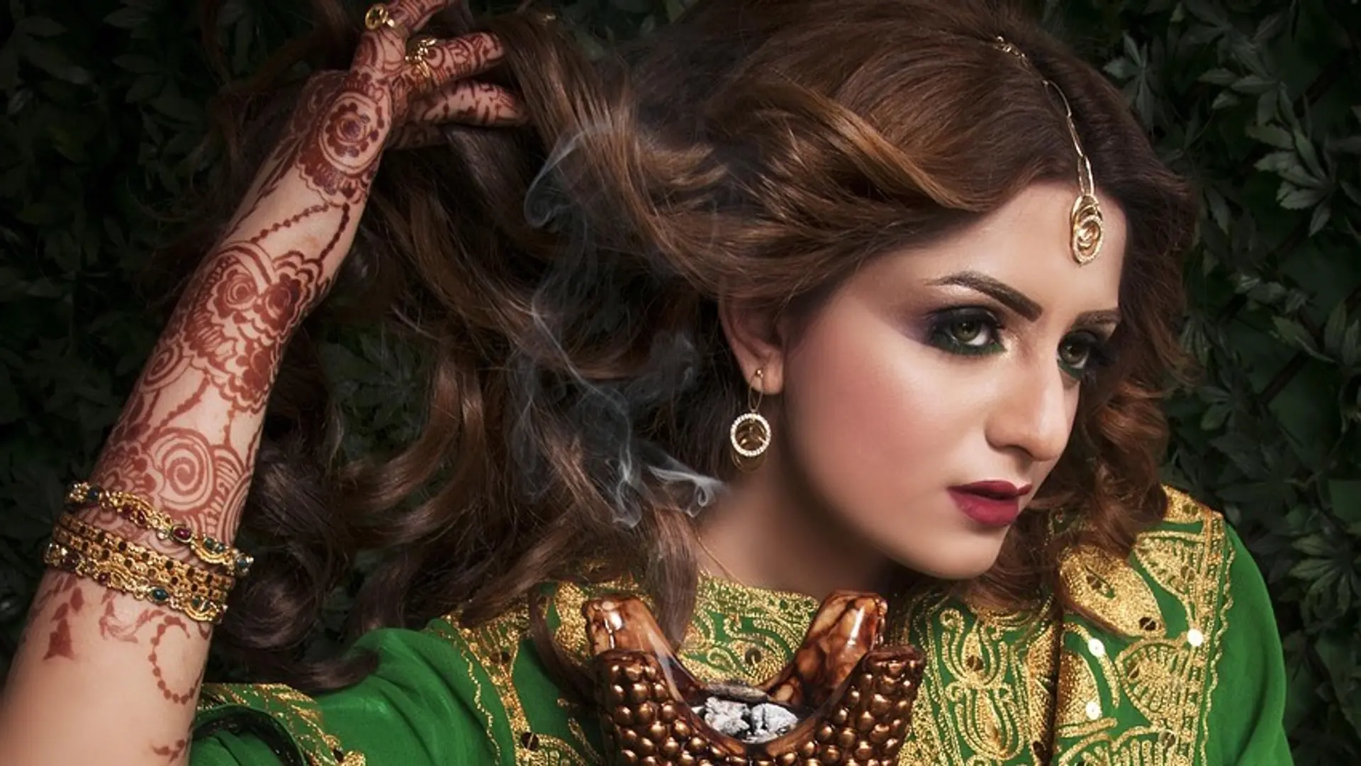 Burnette Indian woman with green and gold dress, arm bracelets, earrings and golden ring at head.