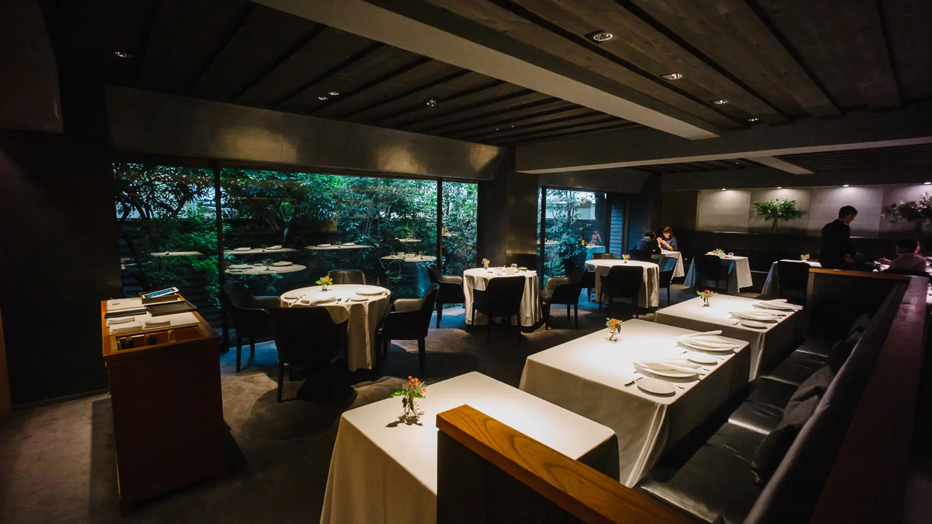 Dark lighted resturant with a contrast window to green nature, dark furniture and white chair at L'Effervescence in Tokyo.