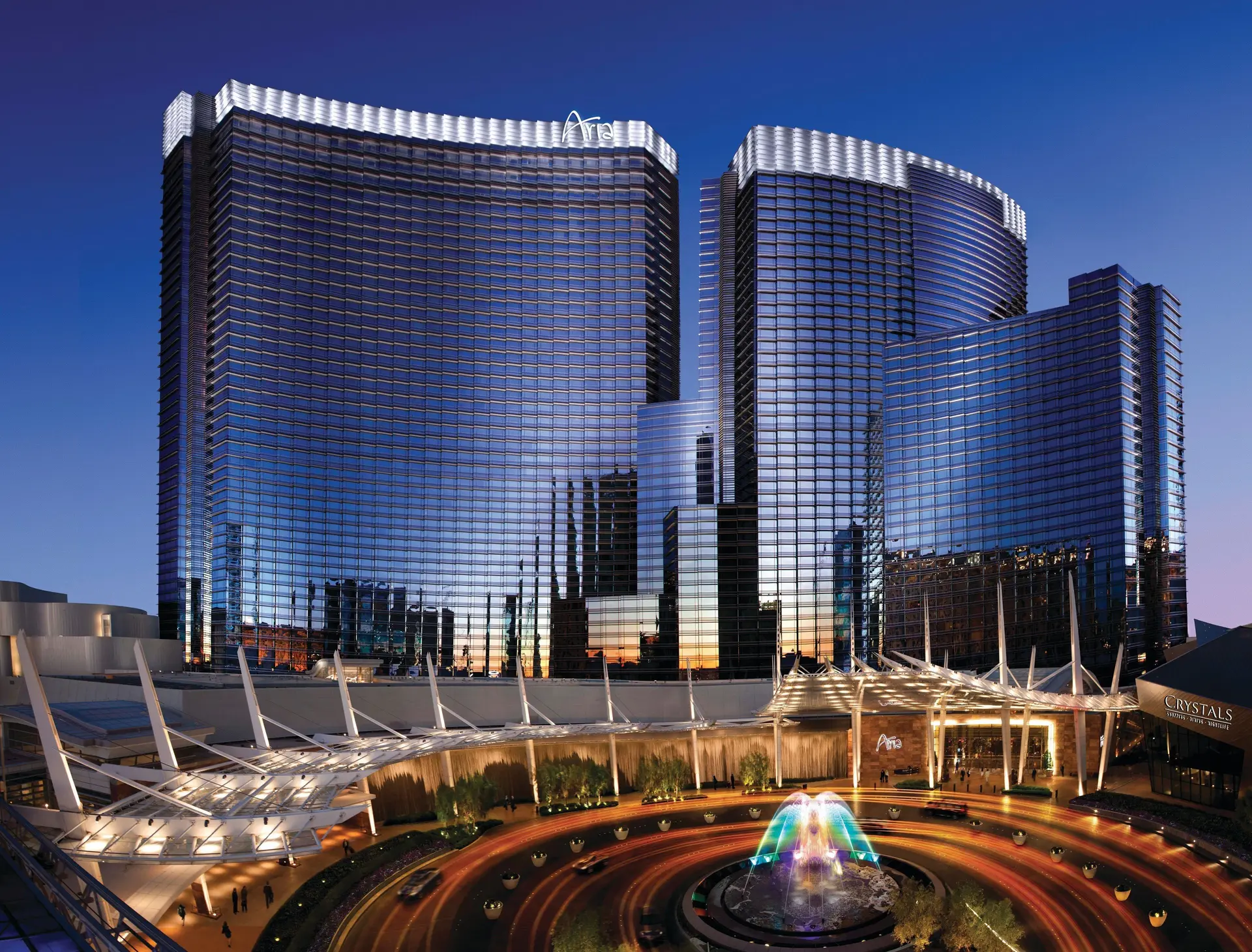 Large dark blue, glass built building, with white at the highest point with text saying: Aria,  and a fountain with colourful waters.