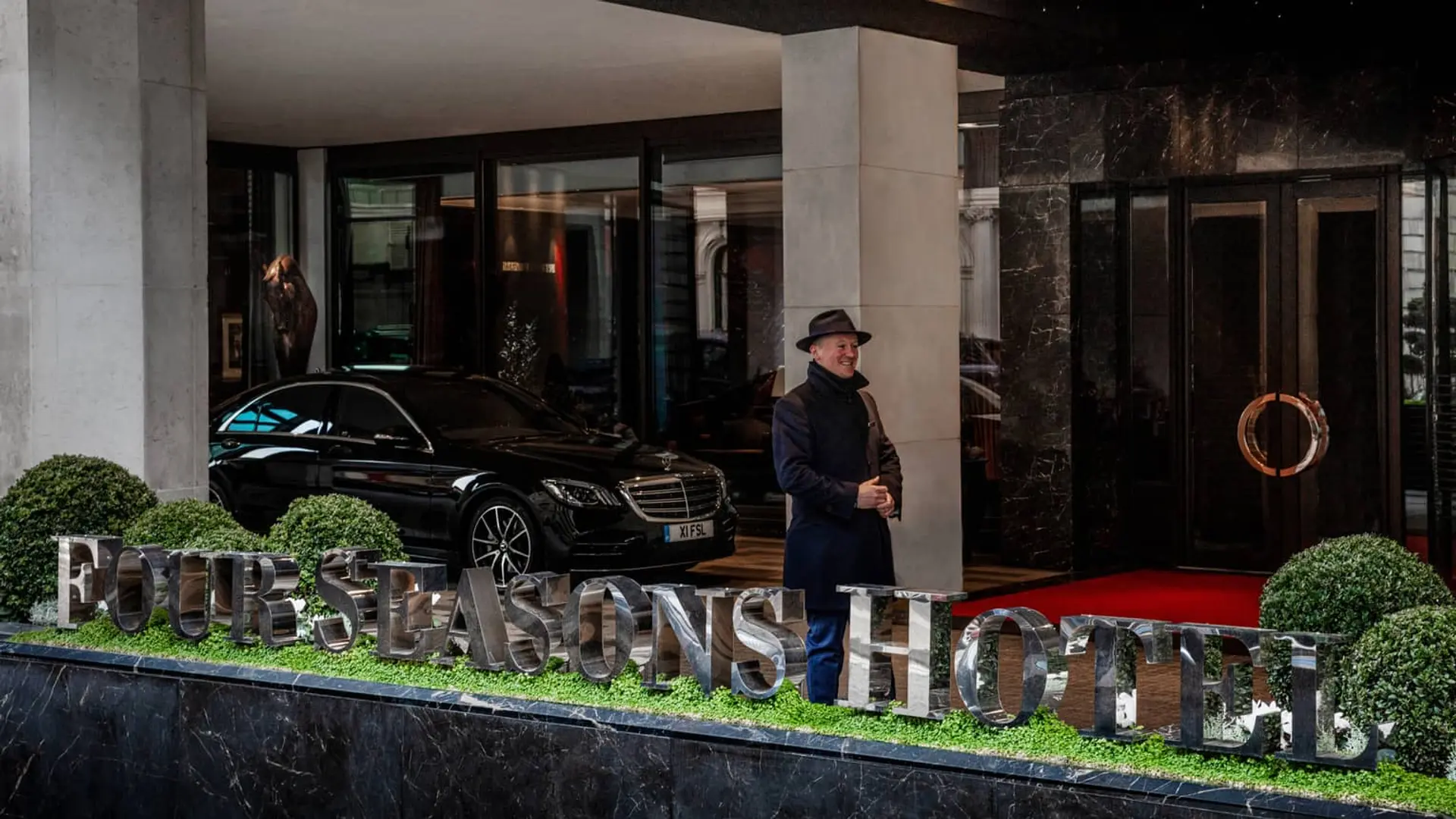 Front entrance of four seasons hotel london at park lane with a black bentley and a man in a coat and a hat.