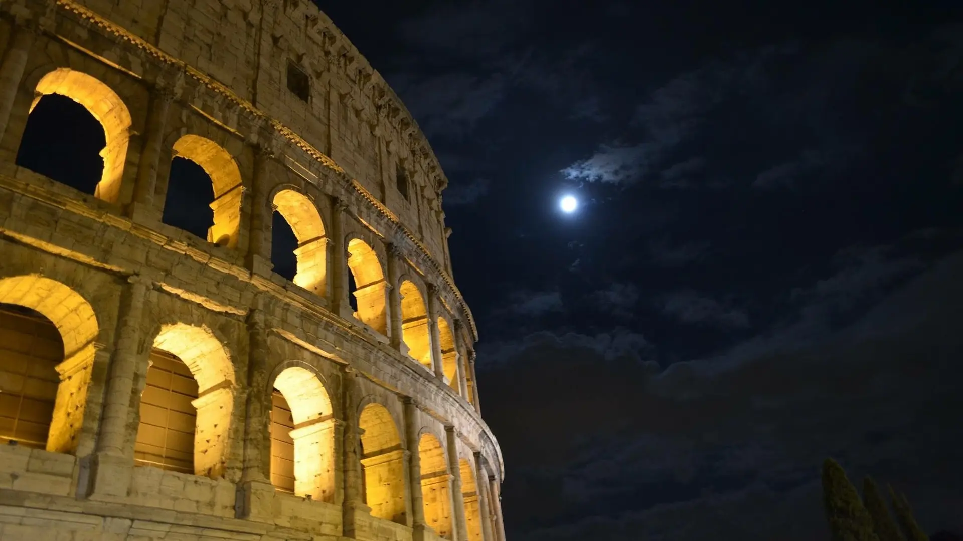 "Exterior view of the Colosseum at night, highlighted by artificial lights that accentuate its grand arches and rugged texture against a dark sky, conveying a sense of historical majesty and enduring architectural significance in Rome's cityscape."