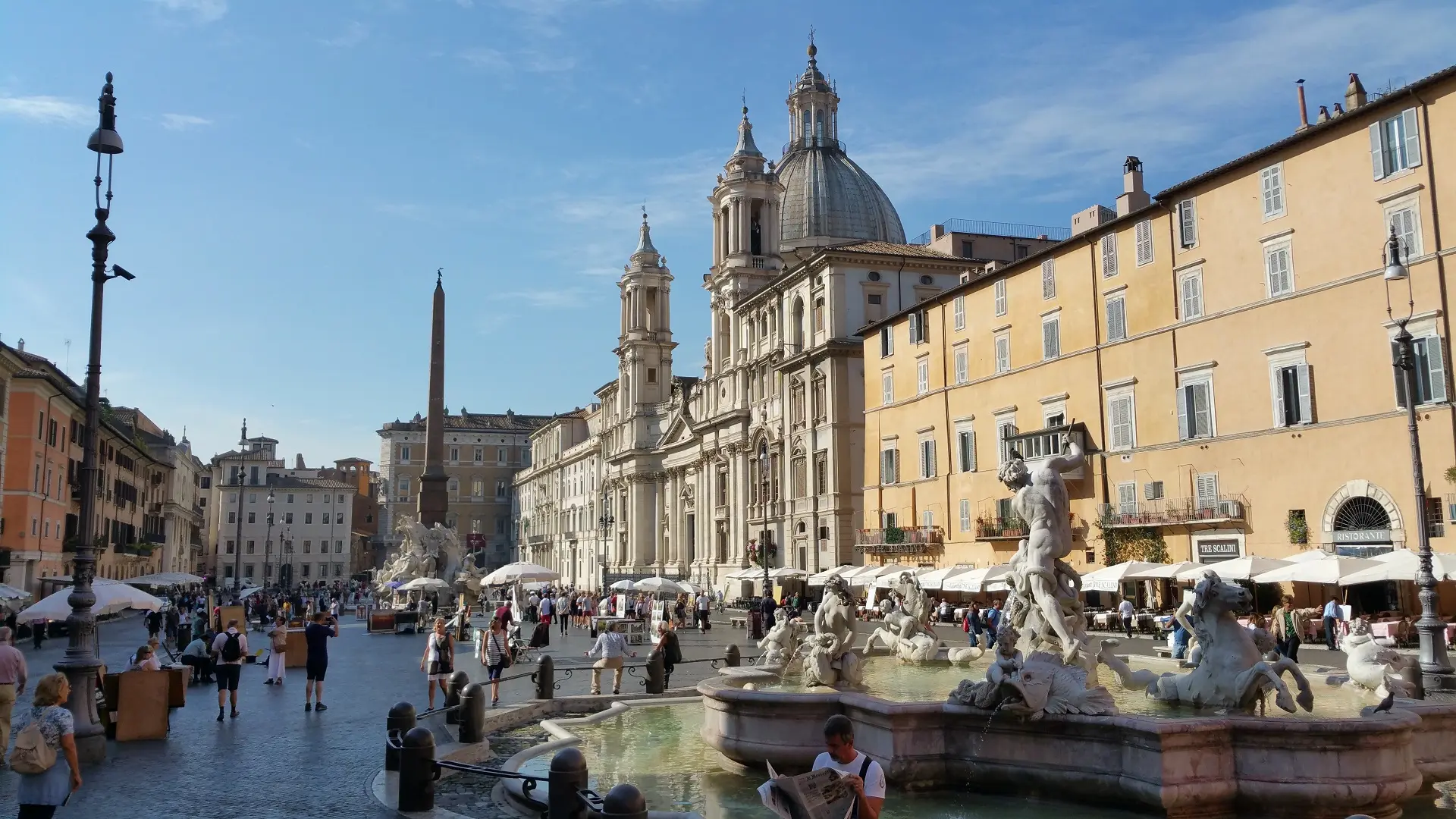 "Photograph of Piazza Navona in Rome, showcasing the bustling square with its famous fountains, Baroque architecture, and outdoor cafes. Visitors and locals are seen strolling, enjoying the vibrant atmosphere under a clear blue sky."