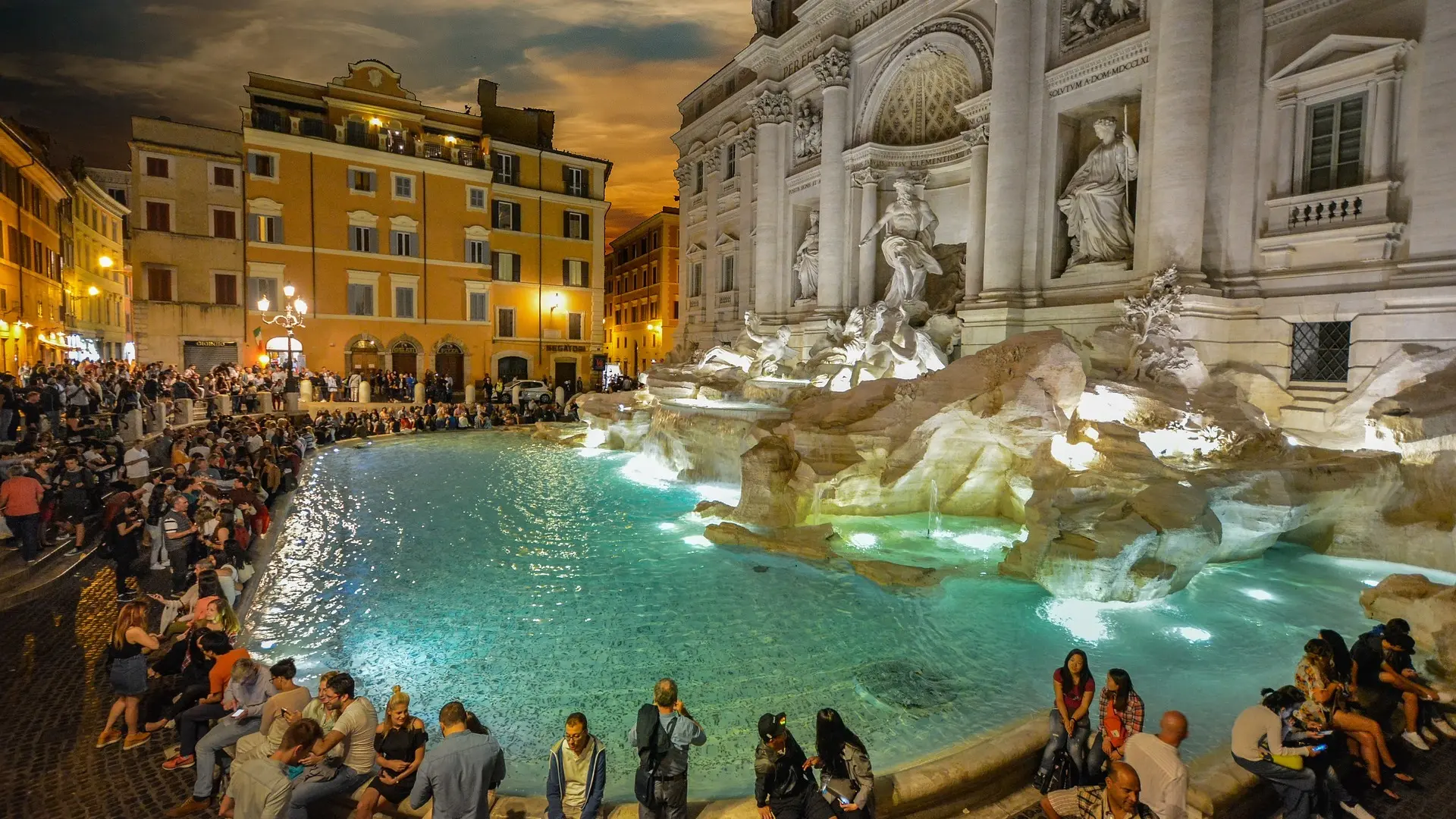 "Nighttime photo of the Trevi Fountain in Rome, showing its detailed sculptures and cascading water bathed in ambient light. Tourists are seen gathered around, some tossing coins, enjoying the fountain's beauty and the lively atmosphere under a starry sky."