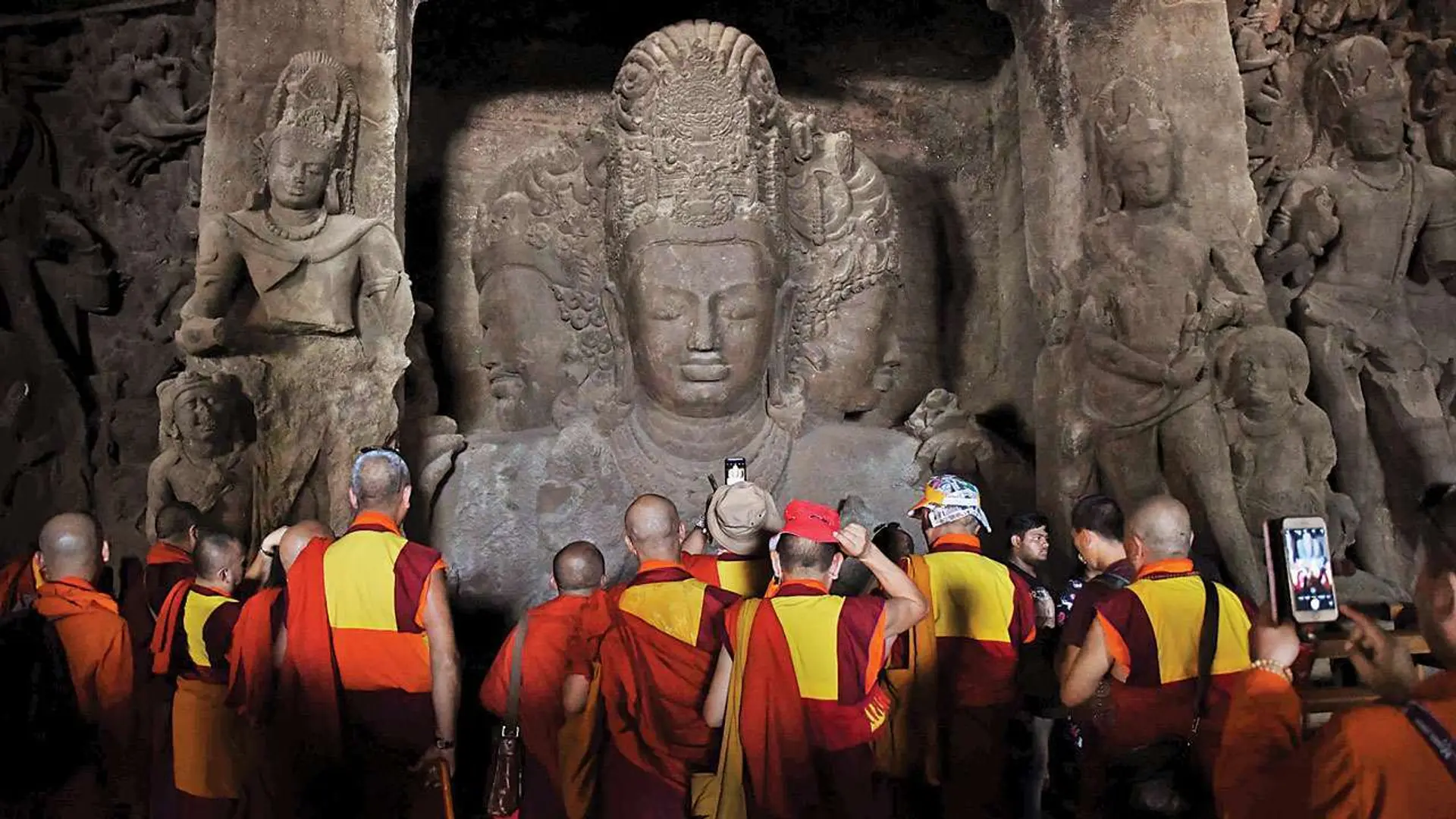 People in red and yellow outfits taking photographs of Buddha like stone engraved statues, this is known as Trimurti in the elephanta caves, Mumbai