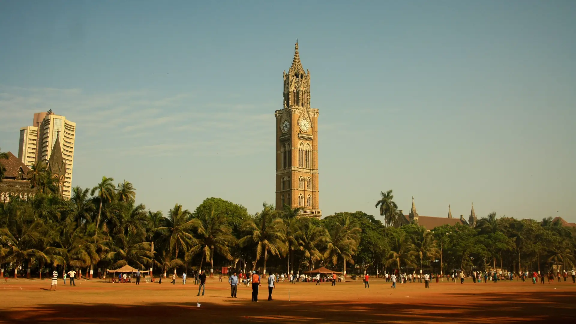 People on a sandy field behind trees and view of Rahq Bhai Tower a tall brick clocktower.