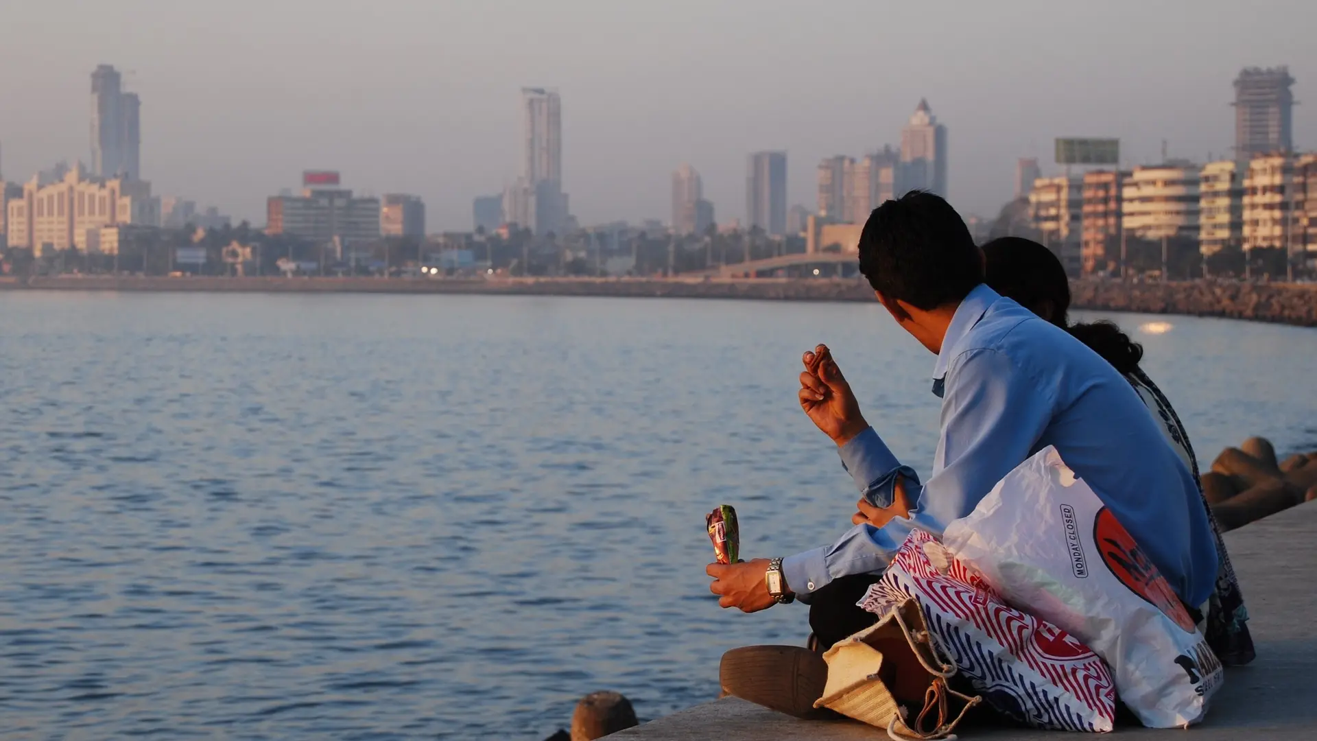 Man in blue shirt with bags sitting beside the shore of Mumbai.