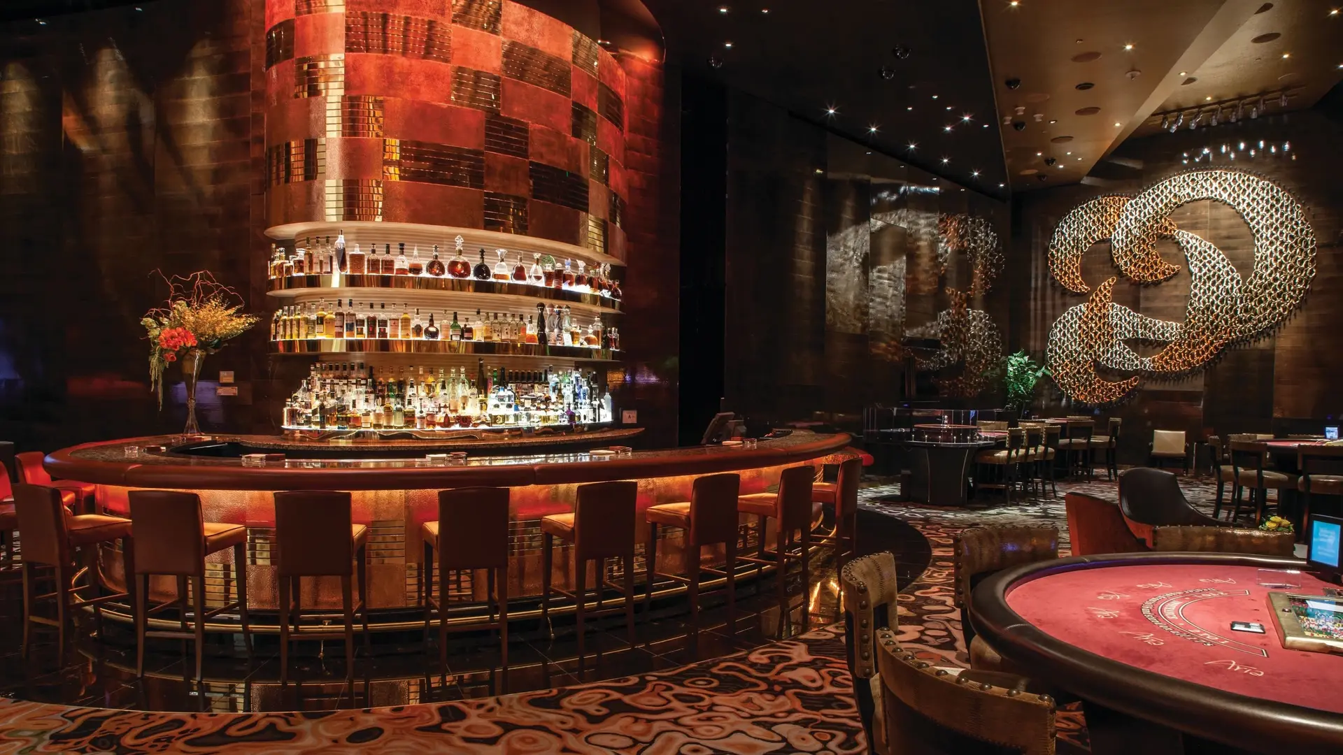 Pub place with tender red ligjting, different drinking options, seating areas, and black wall and poker tables at ARIA Resort å Casino.