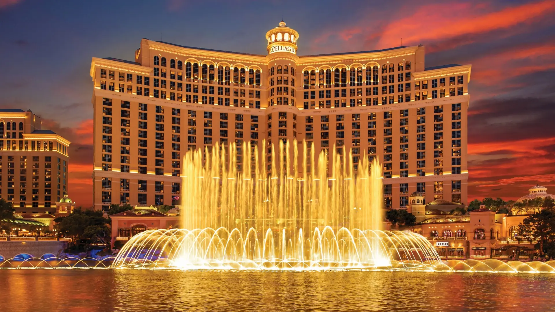 Large fountain with golden coloured water, and wide and tall building behind in a tender yellow colour with a tower in the centre saying: Bellagio. 