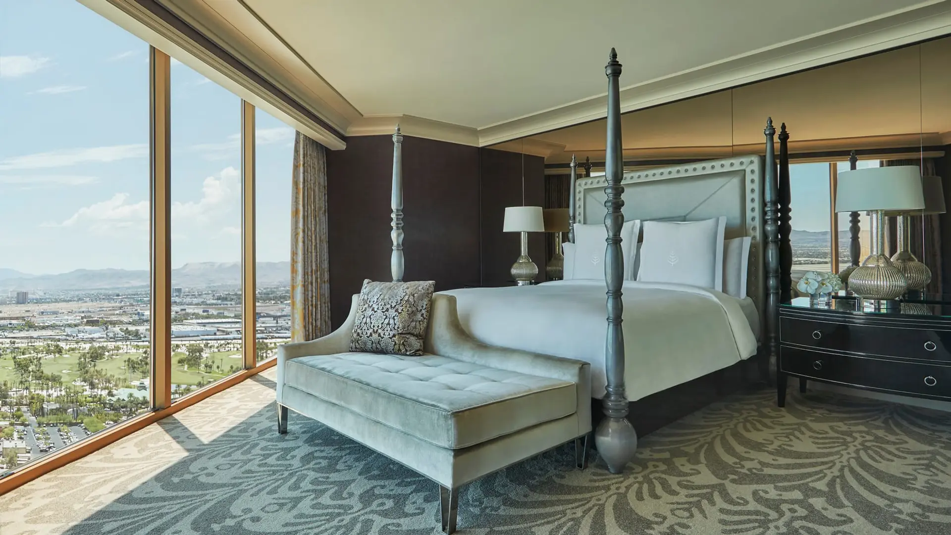 Large window view of Las Vegas, kingsize bed with grey and white decor, sofa in front of bed in grey, walls in a black-ish colour and white roof at The Four Seasons Las Vegas.