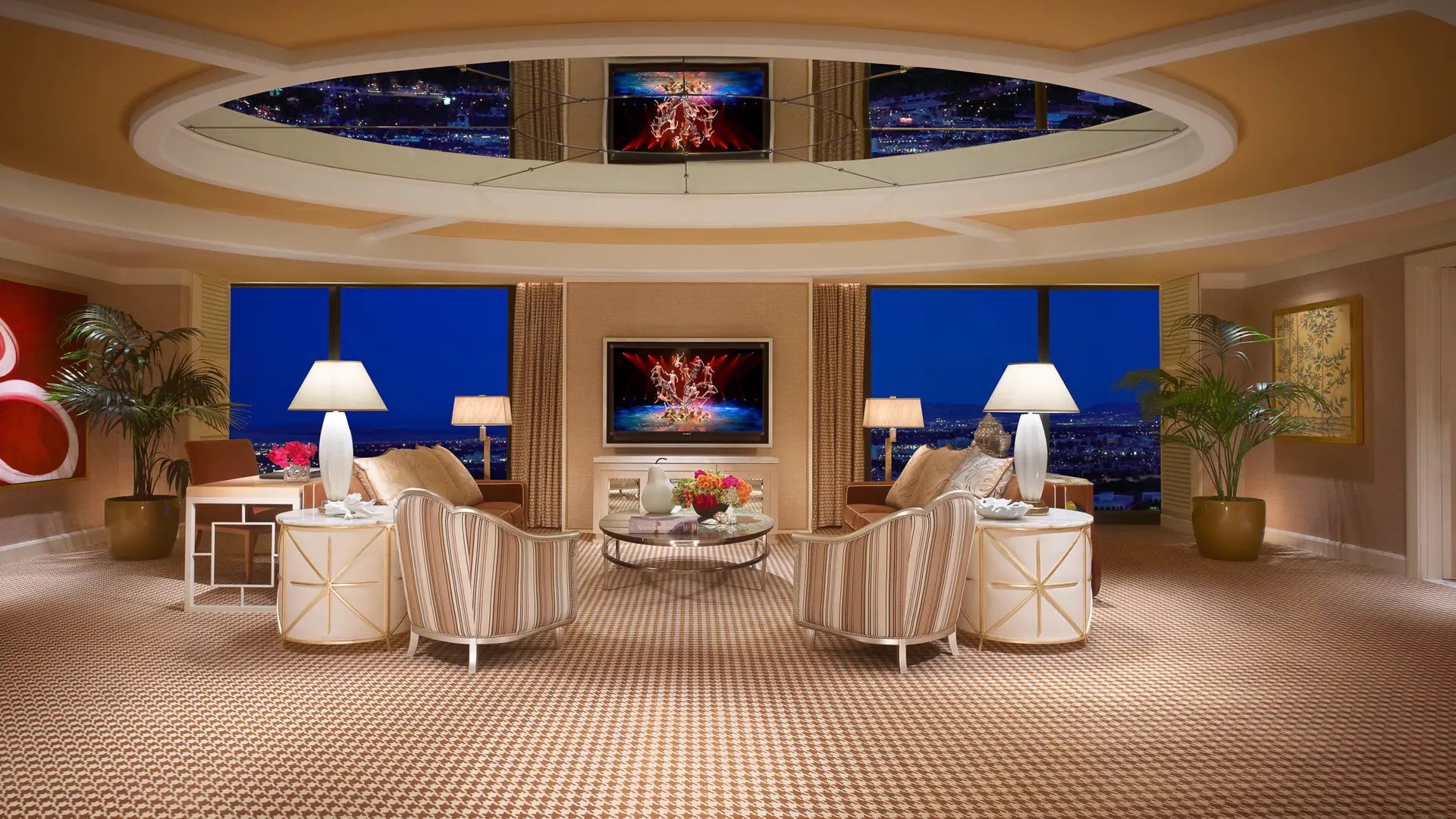 Living room at Wynn Las Vegas å Encore Resort with tv in the centre, two large windows on each side, sofas, and chaior surrounding the tv, lamps, and interior in light white and gold decor.