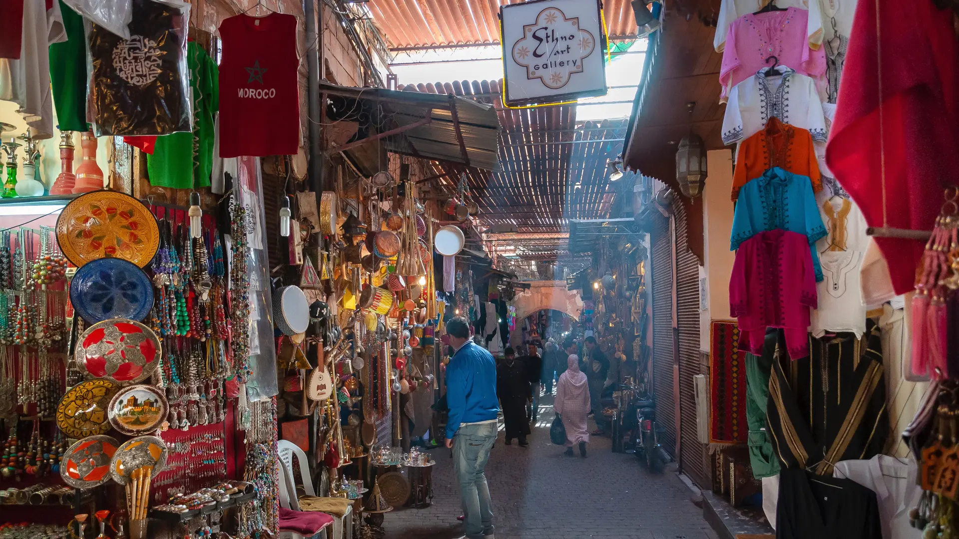 Moroccan market with people walking between. Man in blue jacket.