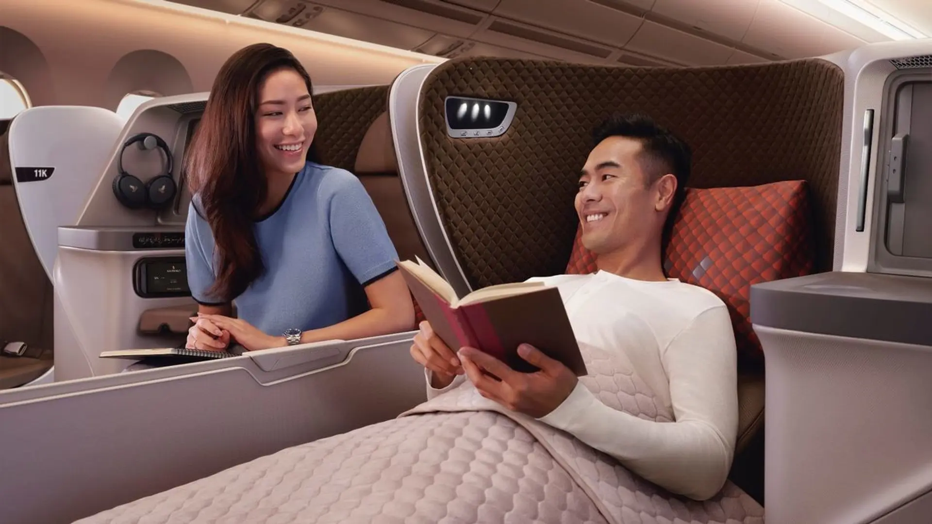 Man laying down on a businessclass flight while reading and smiling at his wife.