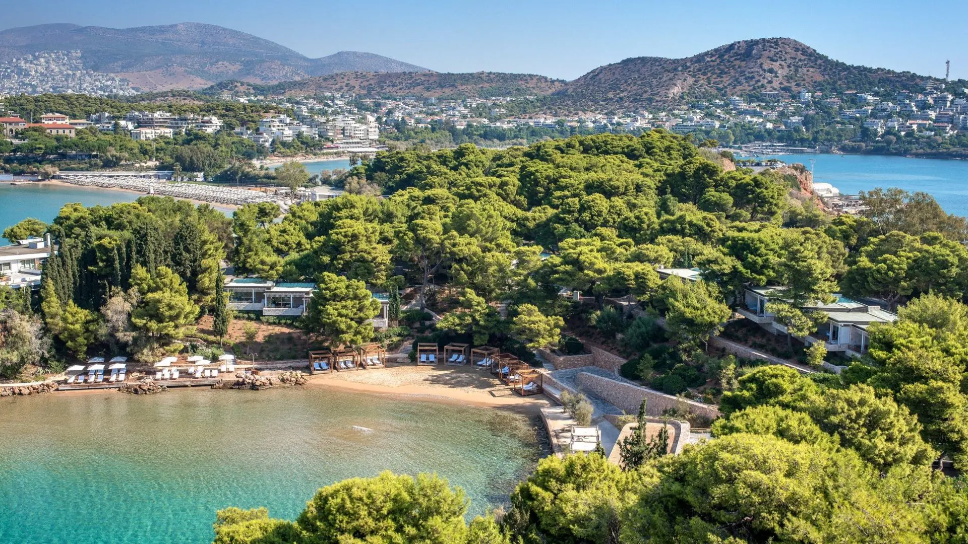 Bird perspective of Four Seasons Astir Palace Hotel in Athens with a lot of nature, petite beach with sunbeds and installed shelter and pastel blue clear water.