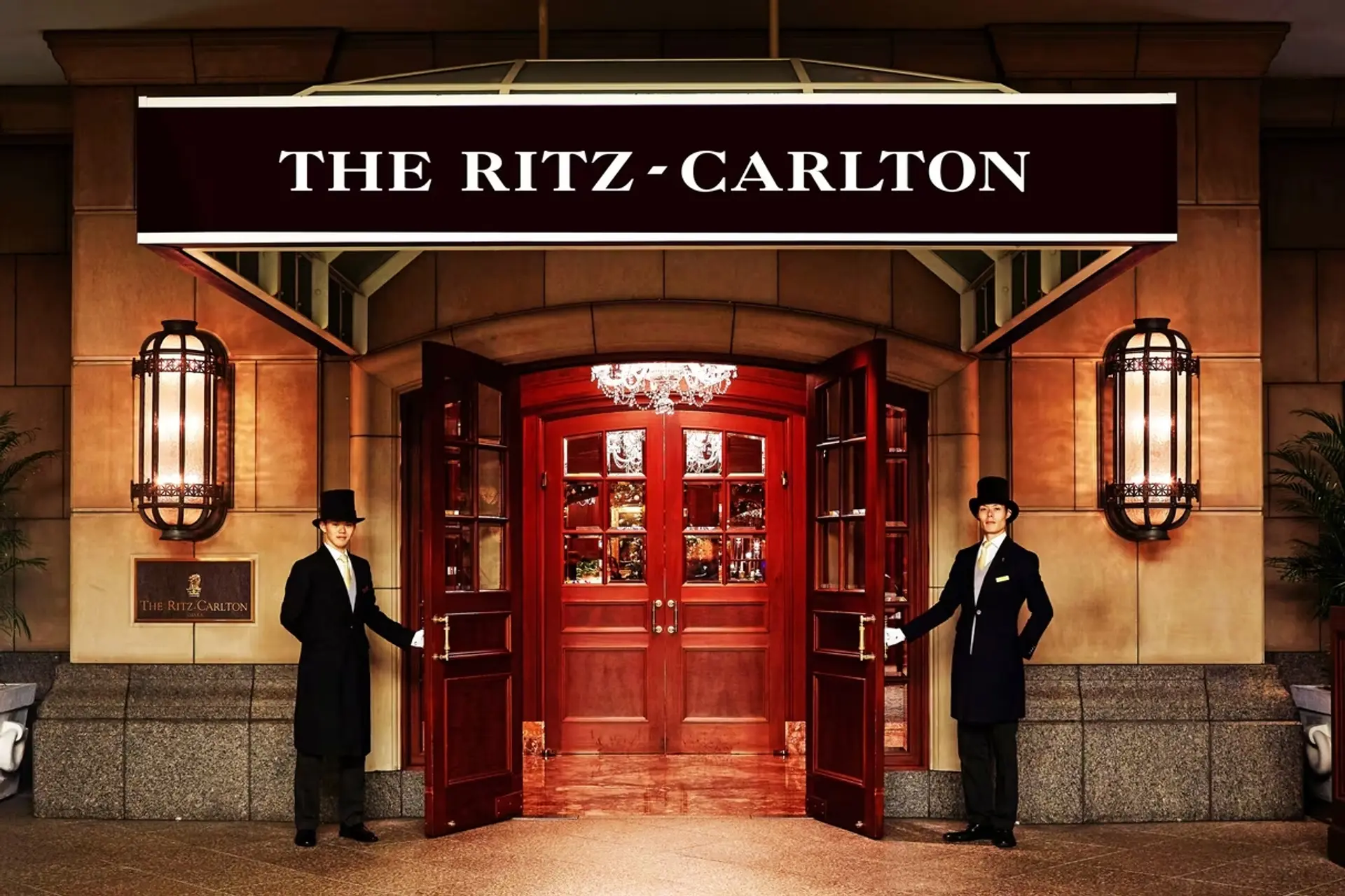 Two men in black uniforms holding the doors open infront of red wooden door and a sign saying: THE RITZ-CARLTON in Osaka.