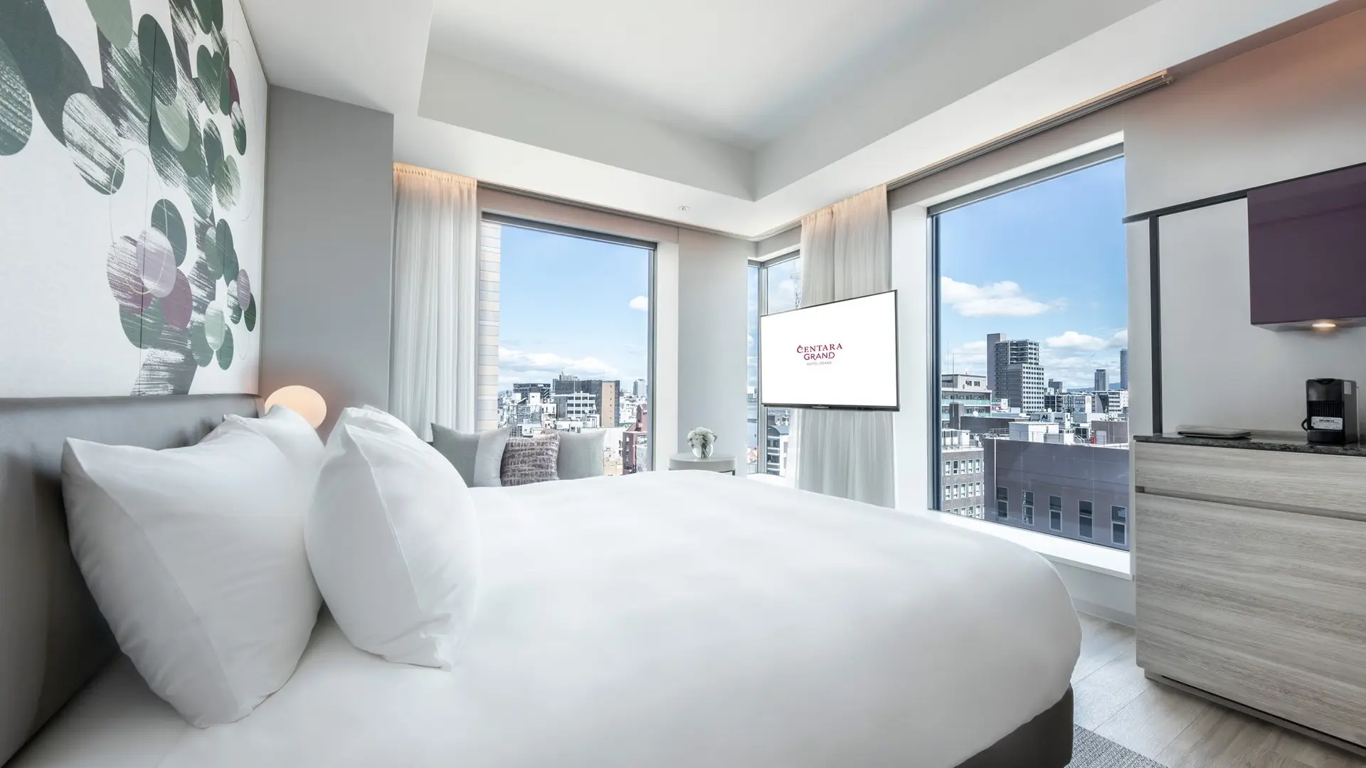 White hotel room with kingsize bed, tv, white modern painting and view to the city.