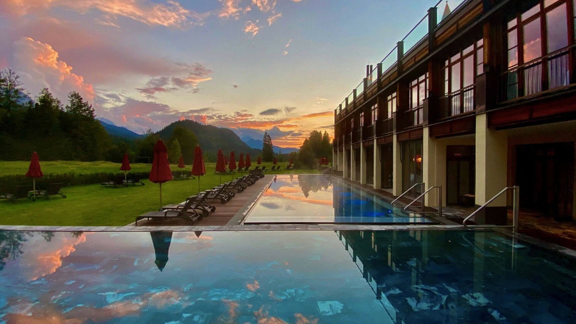 Two outdoor swimming pools with red parasols surrounded by green forests and peaks.