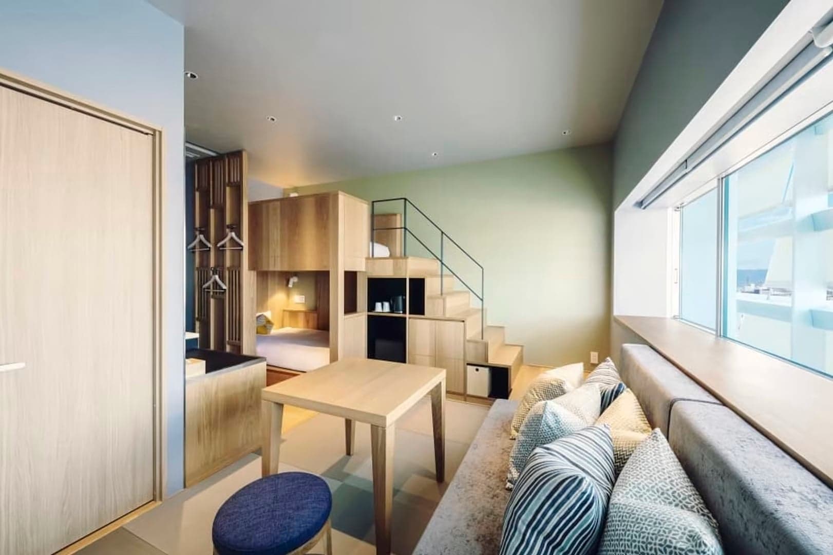 Hotel room in light wood design, seating area in grey, table, small windows, and two beds.