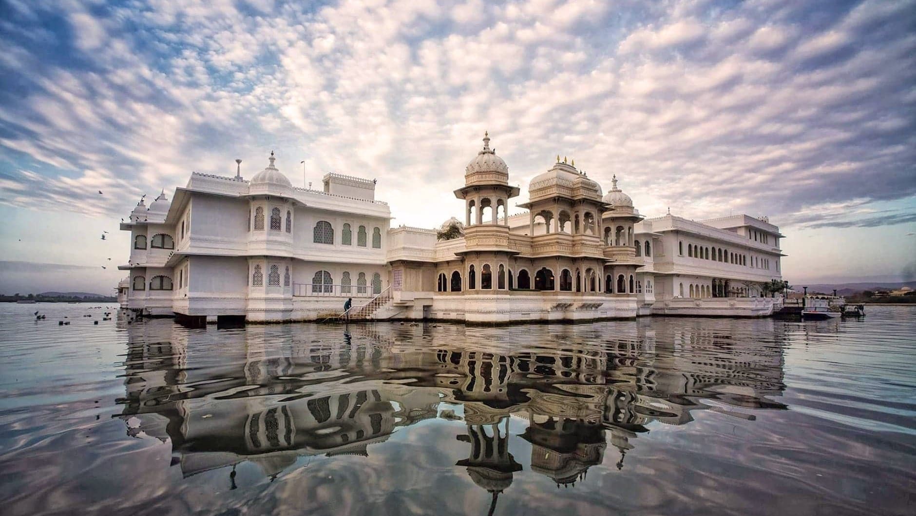 Water view of Taj Lake Palace, a large white palace looking hotel with water sourrounding.