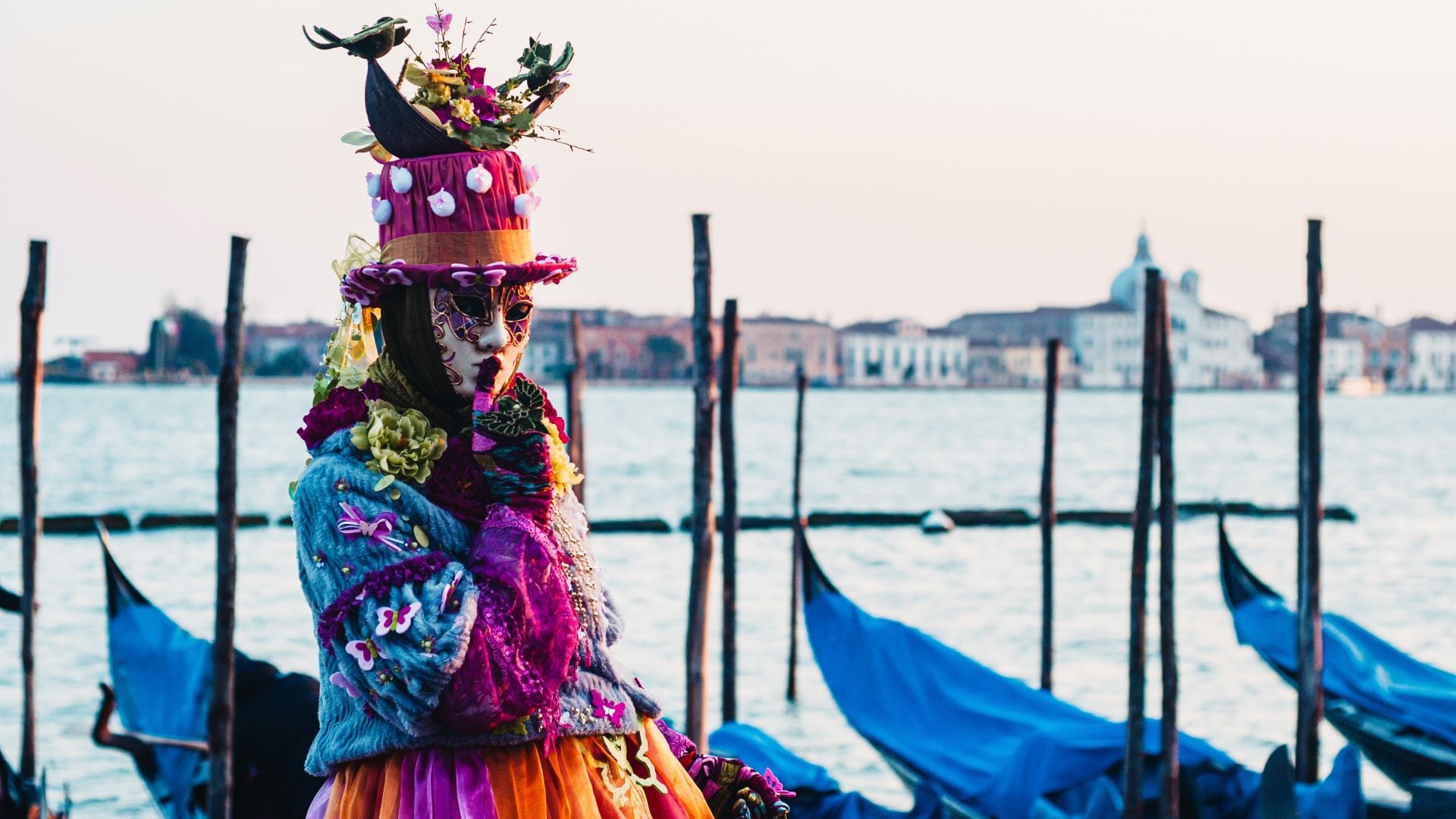 Person in colourful outfit, blue, purple, orange, pink and hat with a mask white and purple making a hush move with his finger in Venice.