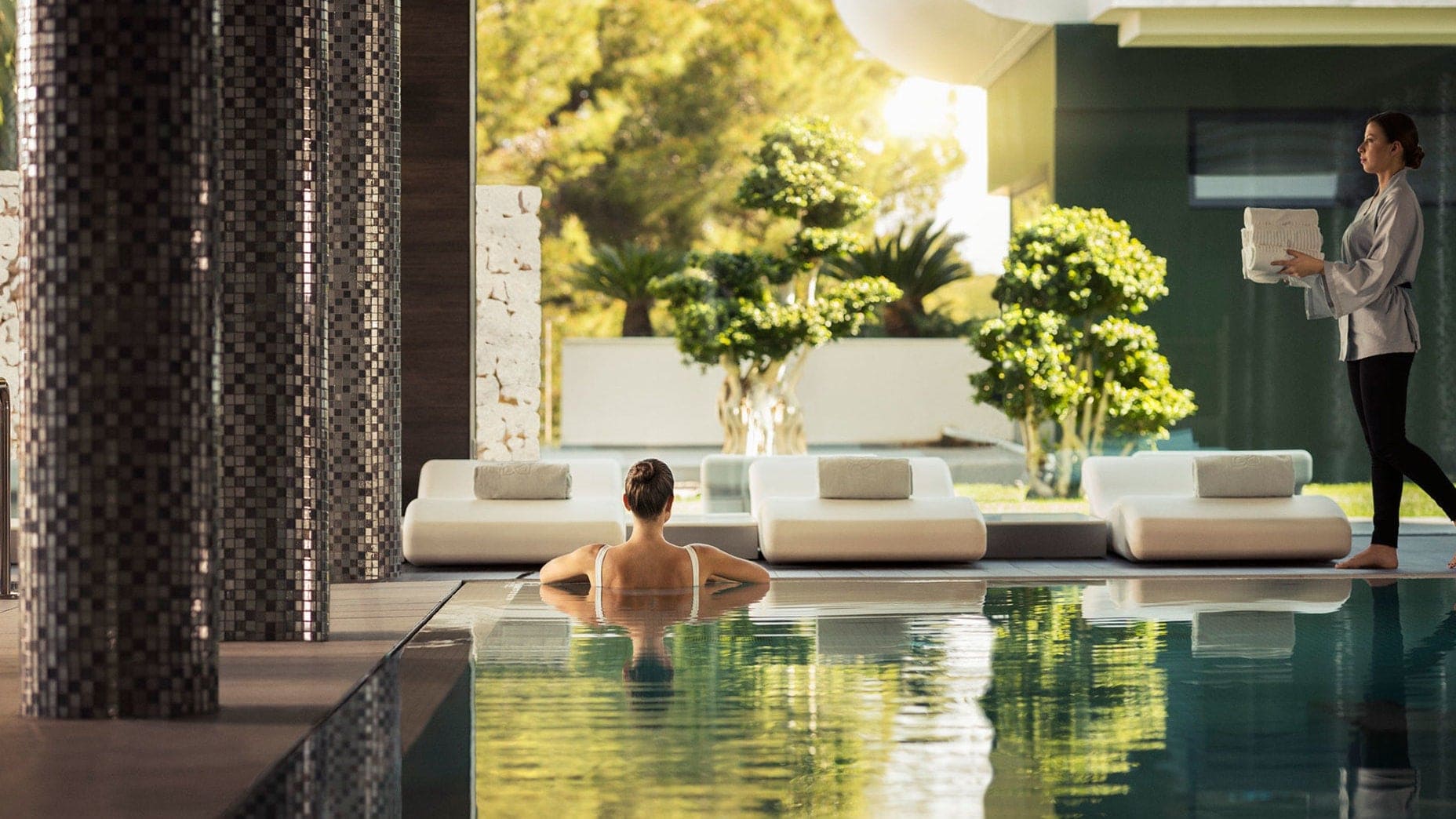 A spa swimming pool with a woman relaxing in the pool and a spa worker walking with towels in her hands.