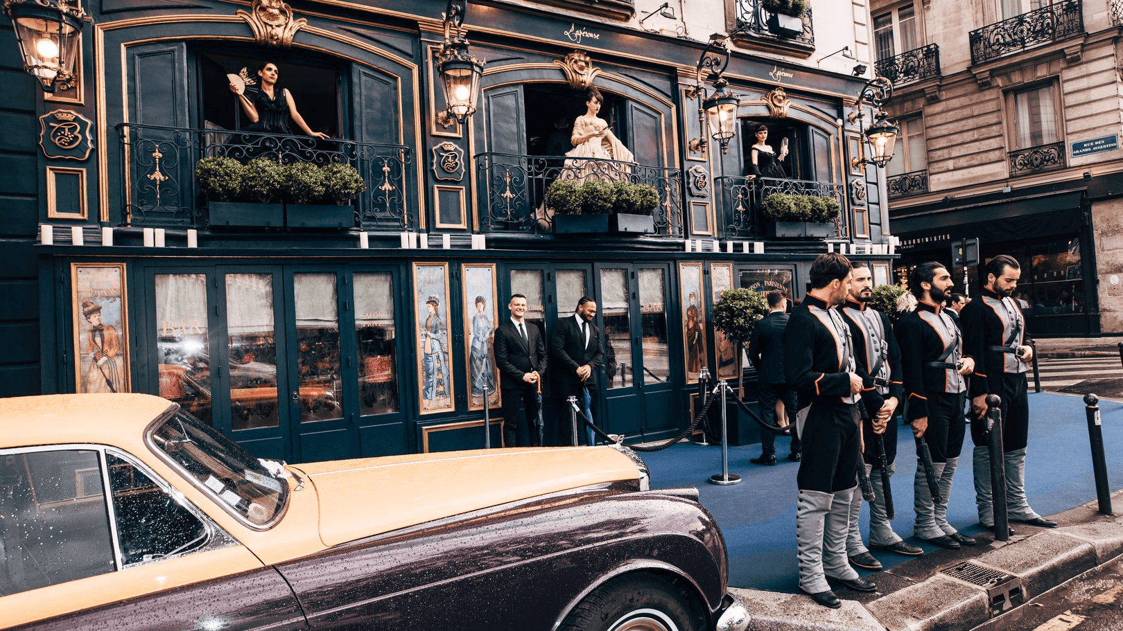 Men in traditional suits standing besides a bentley and a black house.