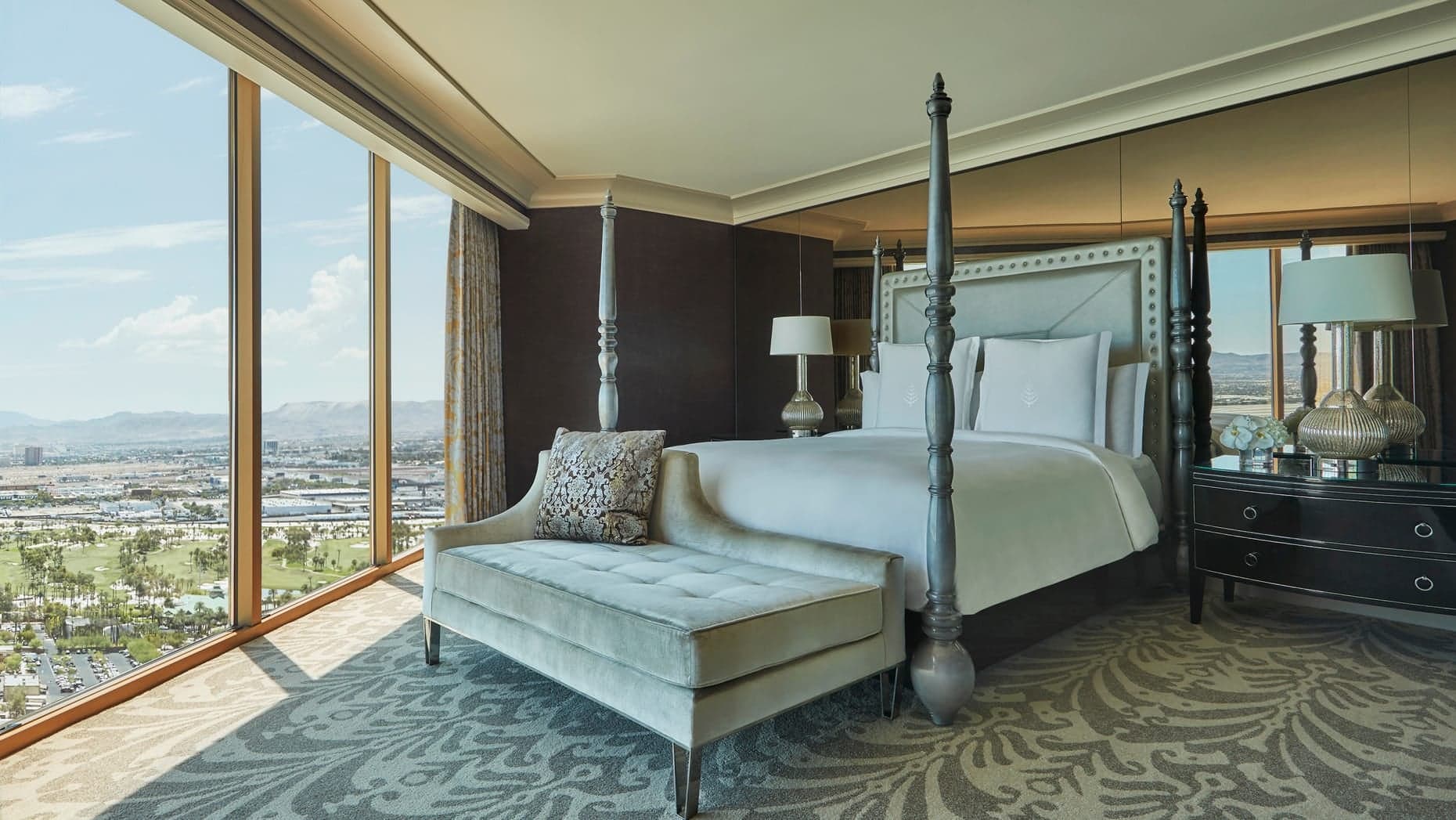 Large window view of Las Vegas, kingsize bed with grey and white decor, sofa in front of bed in grey, walls in a black-ish colour and white roof at The Four Seasons Las Vegas.