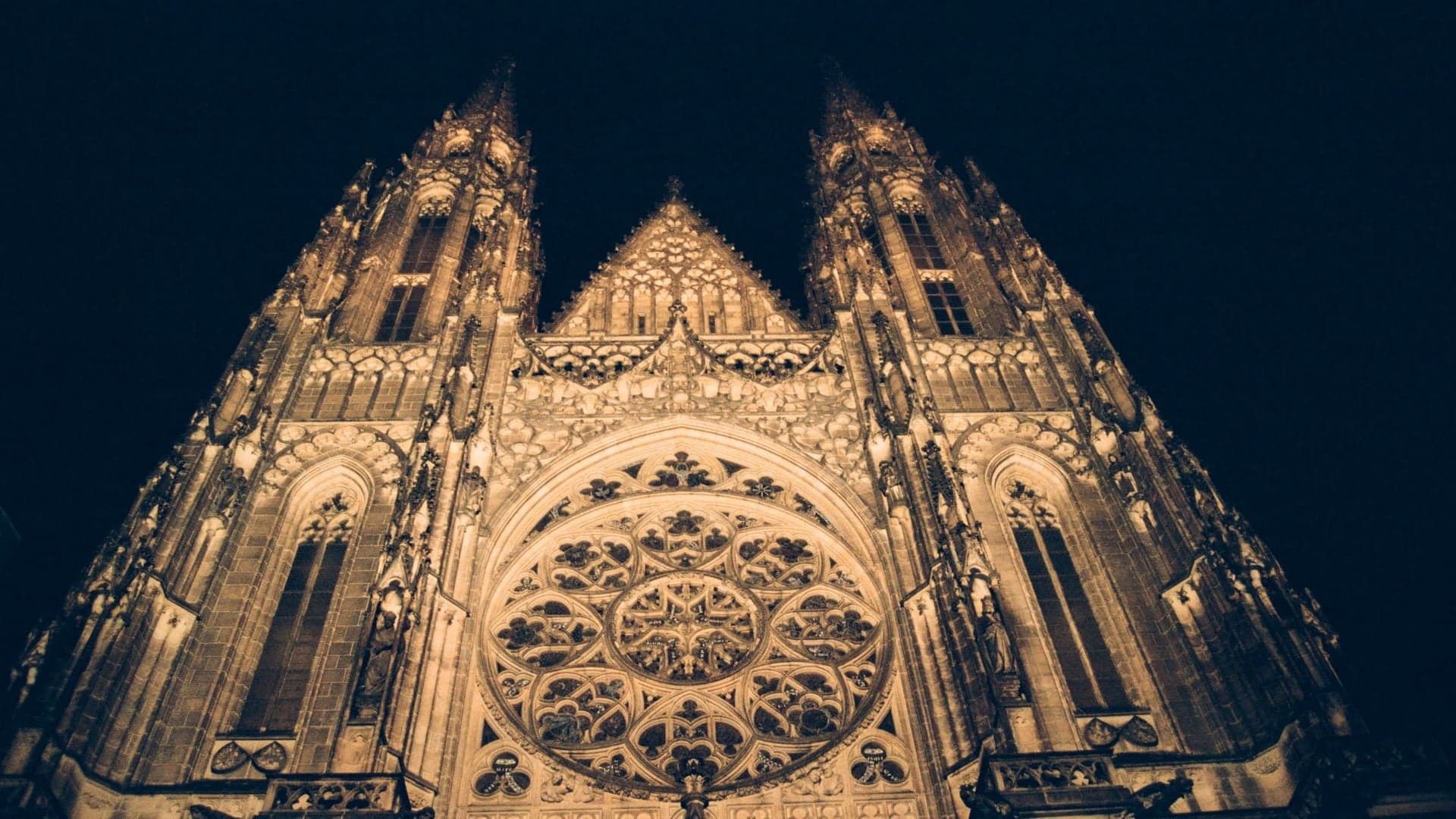 Frog perspective of St Vitus Cathedral, a large symmetrical building in gold lightning and to towers on each side with black backround. In Prague, the Czech Republic.