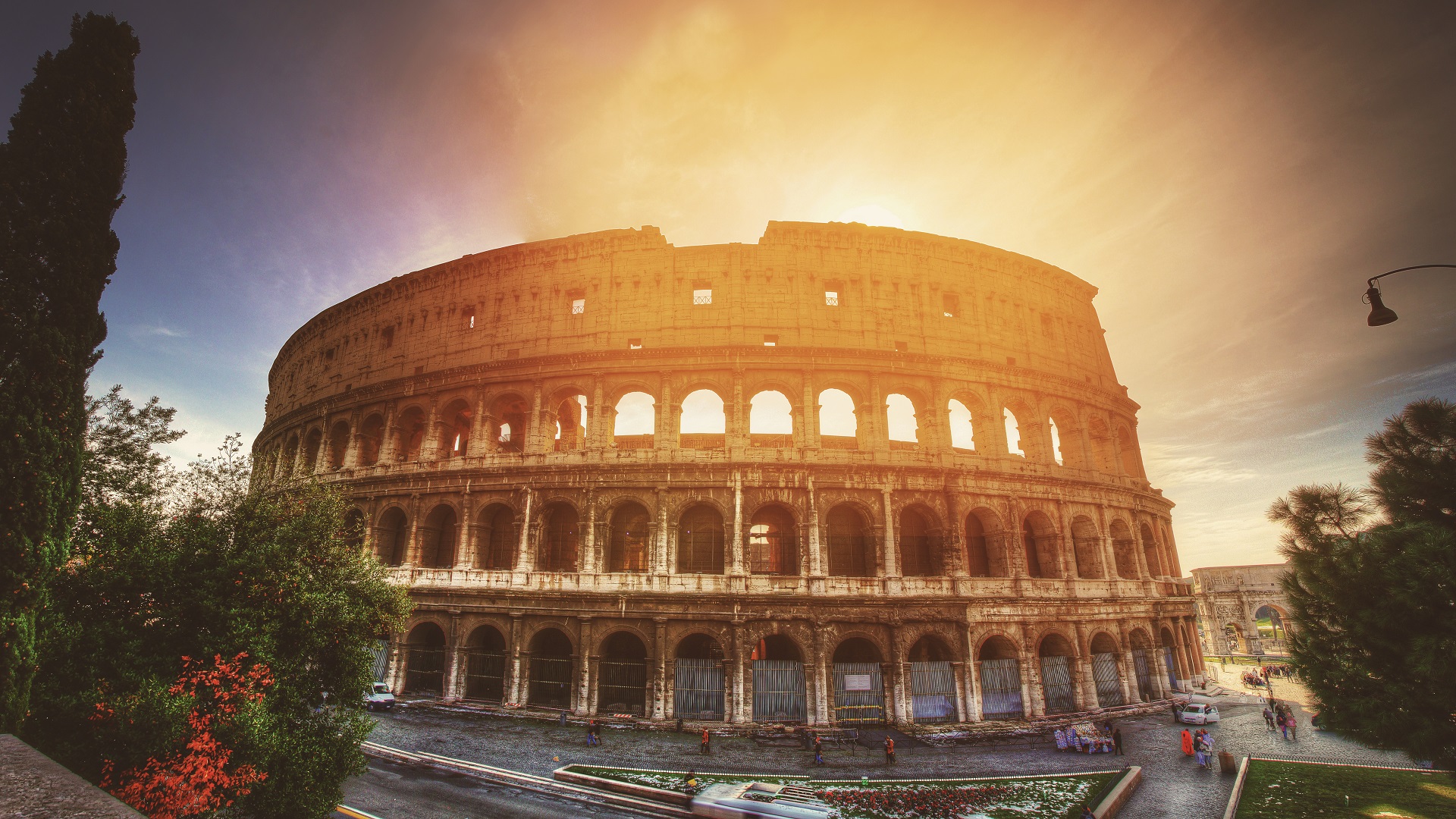 "Photo of the Colosseum in Rome during sunset, with the iconic ancient amphitheater bathed in the warm glow of the setting sun, casting long shadows and highlighting its historic arches and weathered stone structure against a vibrant, colorful sky."
