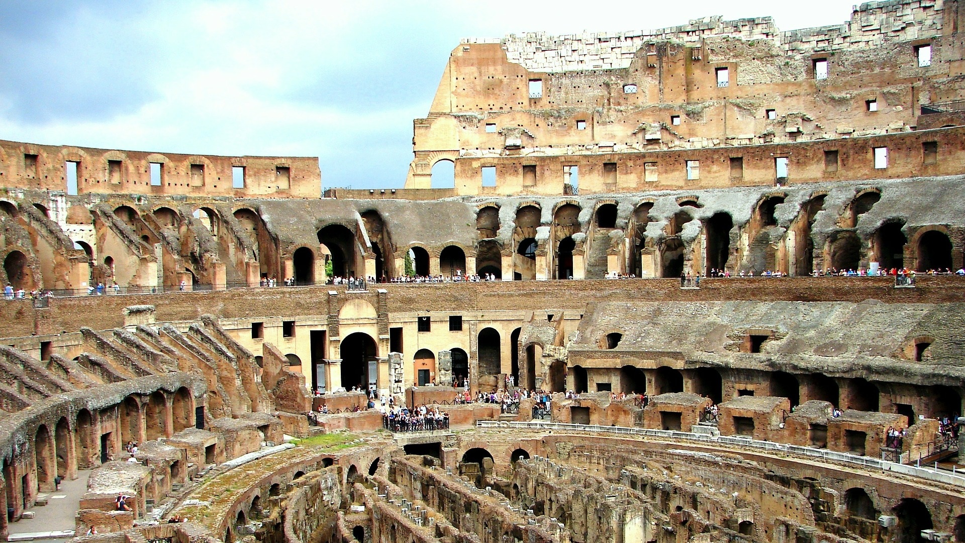 "Image showing the interior of the Colosseum in Rome during daytime, bustling with tourists exploring the ancient arena. Sunlight streams in, illuminating the aged stone walls and arches, highlighting the grandeur and scale of this historic monument."