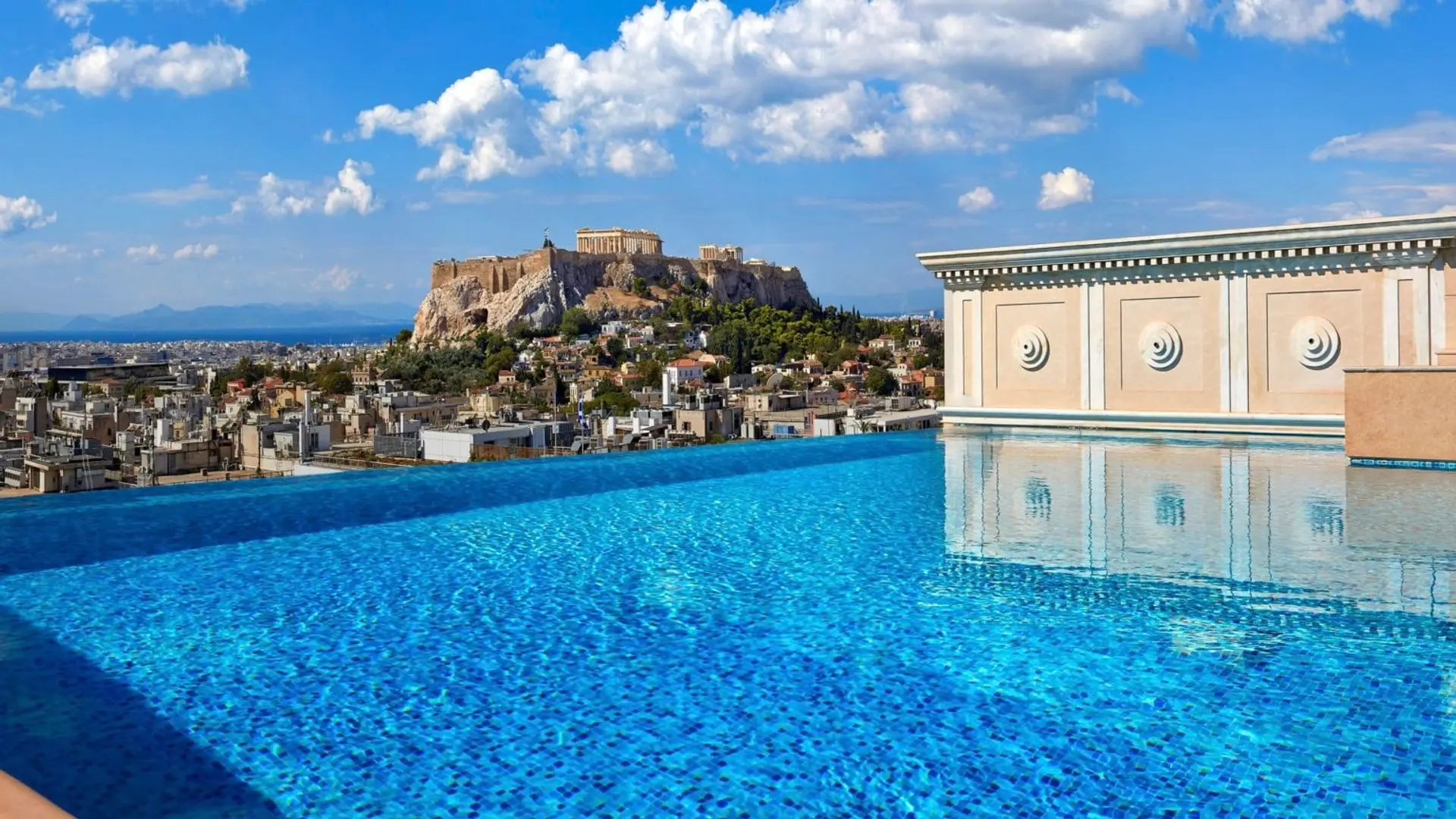 Infinity pool with clear blue water, and a insane view of nature, the city of Athens and a ancient building on top of mountain.
