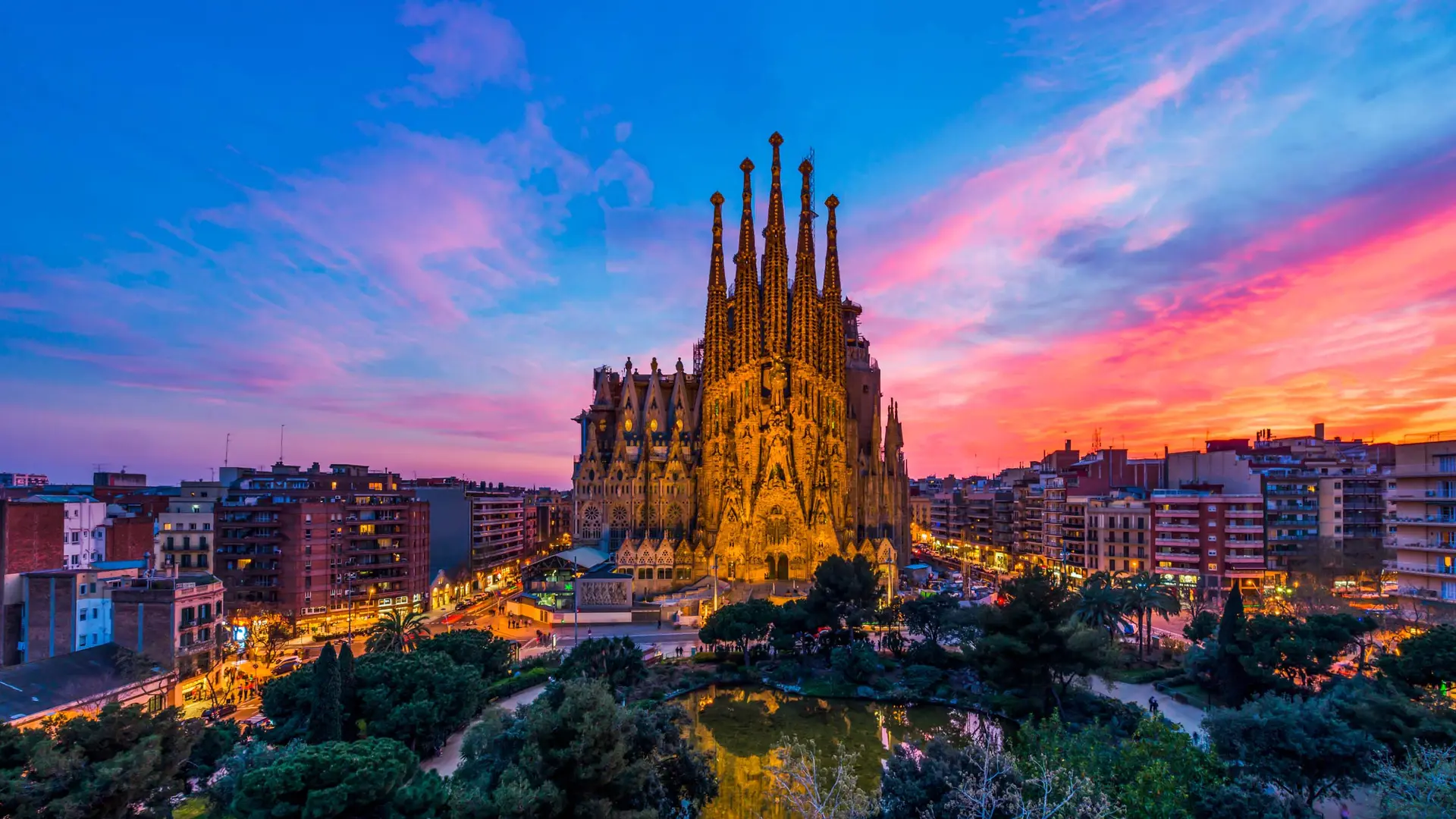 View of the iconic The Sagrada Familia with a tall tower, beautiful sunset, and large park with water and nature in Barcelona, Spain.