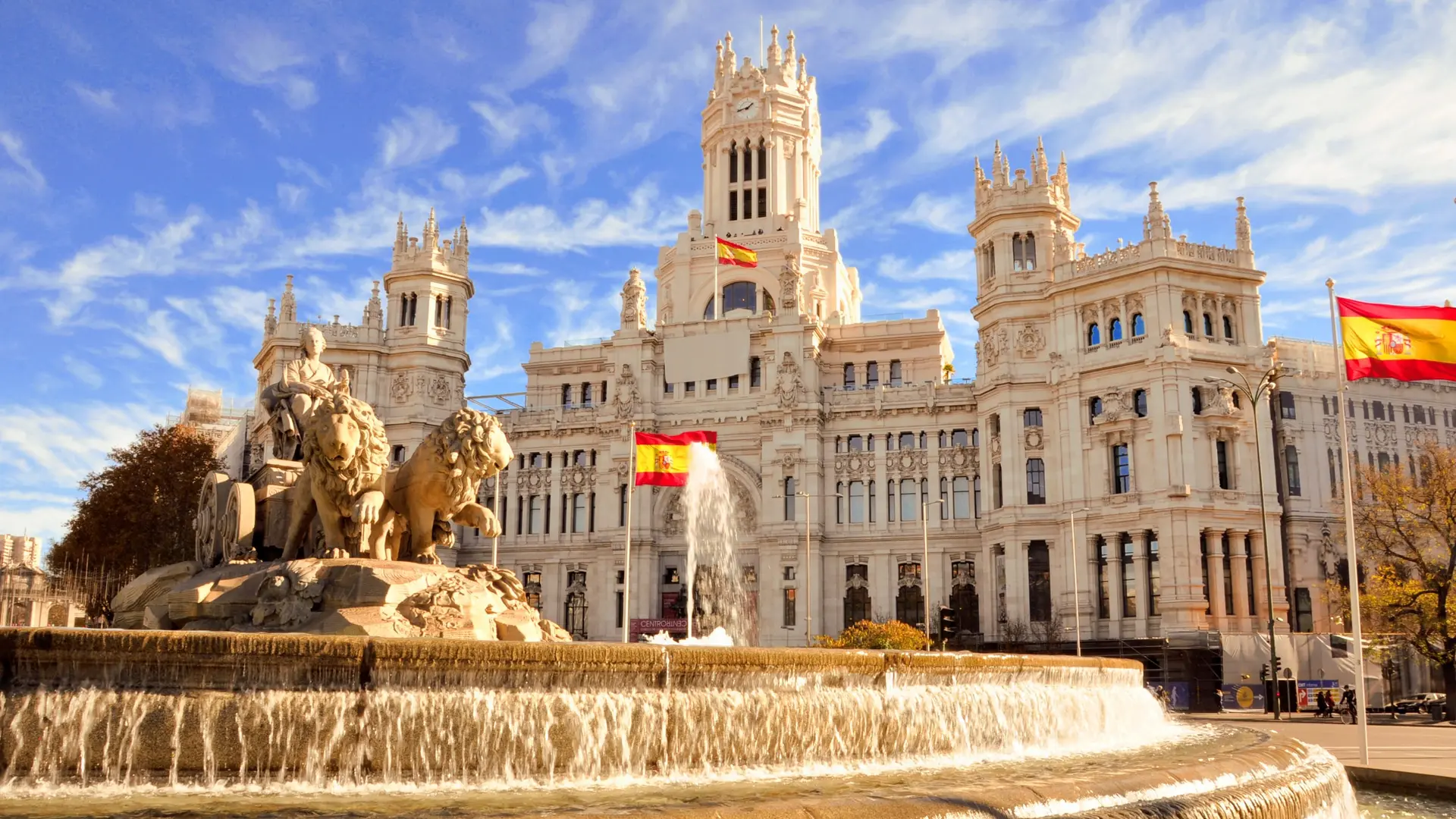 View of the cibeles fountain and large pastel sand white colioured castle behind and a lot of spanish flags.