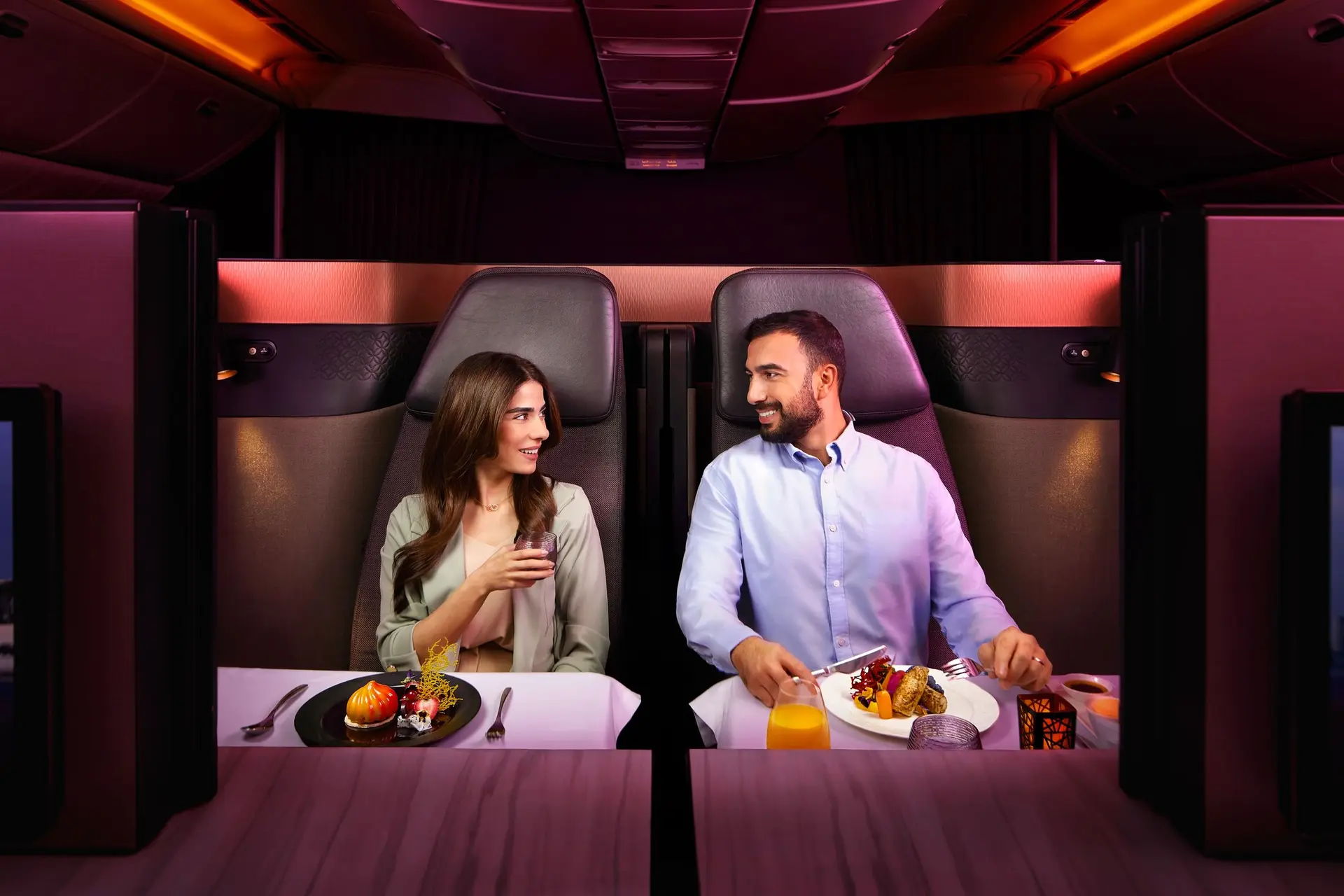 Happy couple sitting on double seats on a businessclass flight smiling at eachother while enjoying a meal.