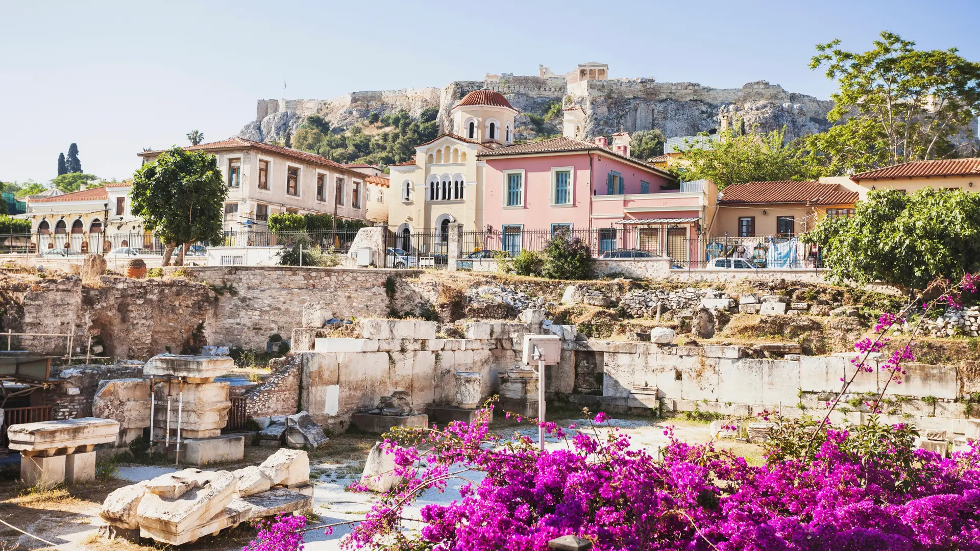 A place in athens with purple flowers in the foreground and some houses with a cliff in the background.
