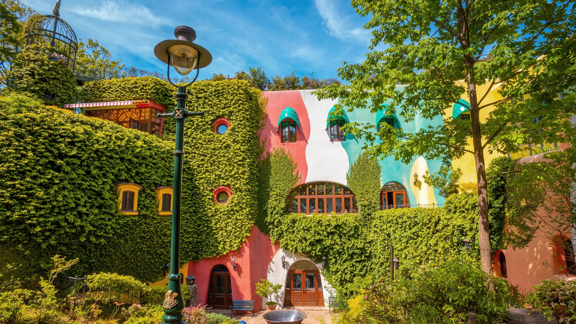 Pink, white, pastel blue and yellow building with bushes covering 50% at Ghibli Museum.