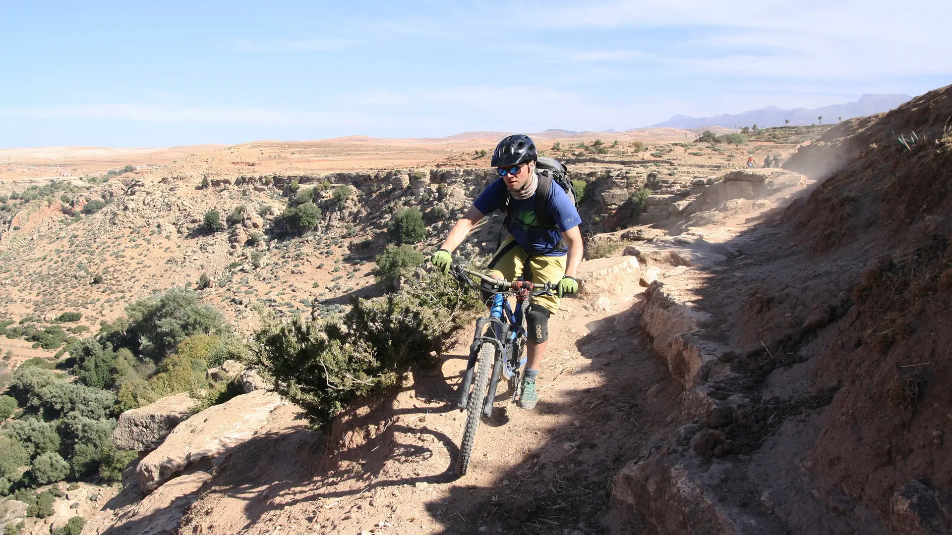 Guy riding a terrain bike on mountains wearing sunglasses. a blue shirt and a black helmet.